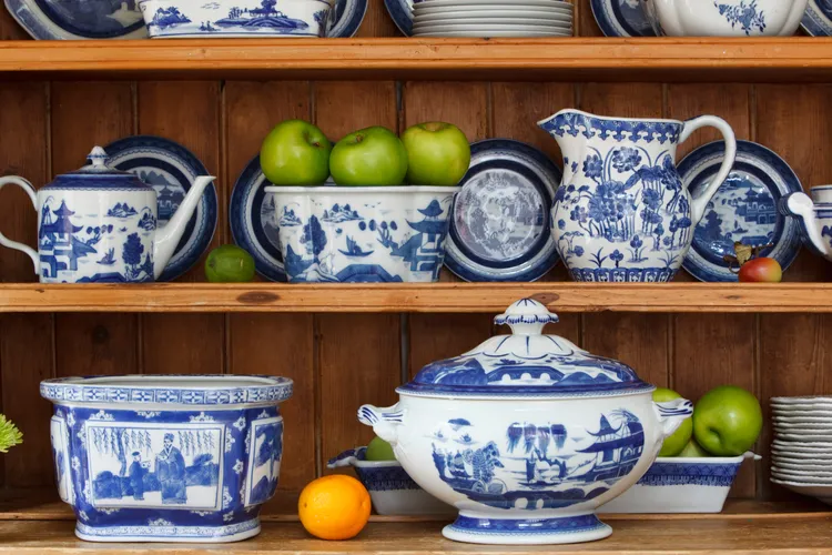 Blue and white china dishes and pitchers are arranged on wooden shelves, with green apples and an orange placed among the dishes.