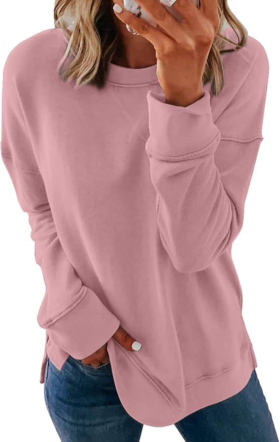 A person wearing a loose-fitting, light pink sweatshirt and blue jeans, holding a phone with one hand and the other hand in a pocket.