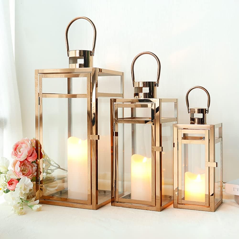 Three gold-framed glass lanterns of varying sizes with lit candles inside are arranged next to a small bouquet of pink and white flowers.