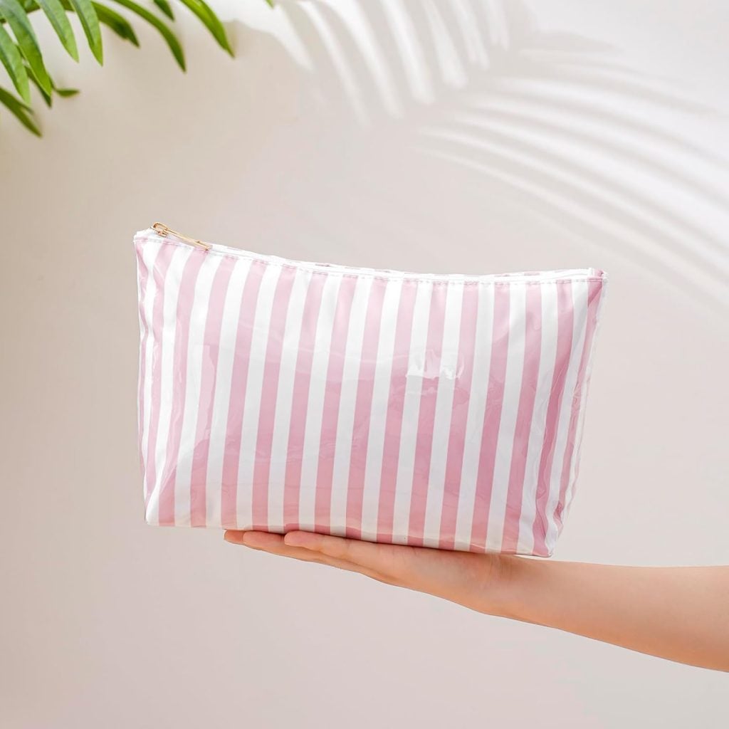 A hand holds a pink and white striped zippered pouch against a light background with plant shadows.