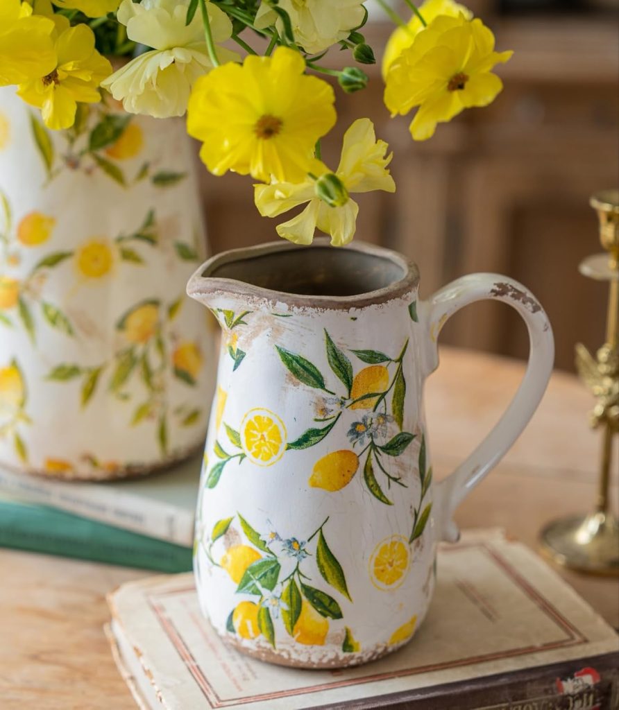 White ceramic pitcher with a lemon pattern holds yellow flowers, placed on stacked books with a matching pitcher and candlestick in the background.