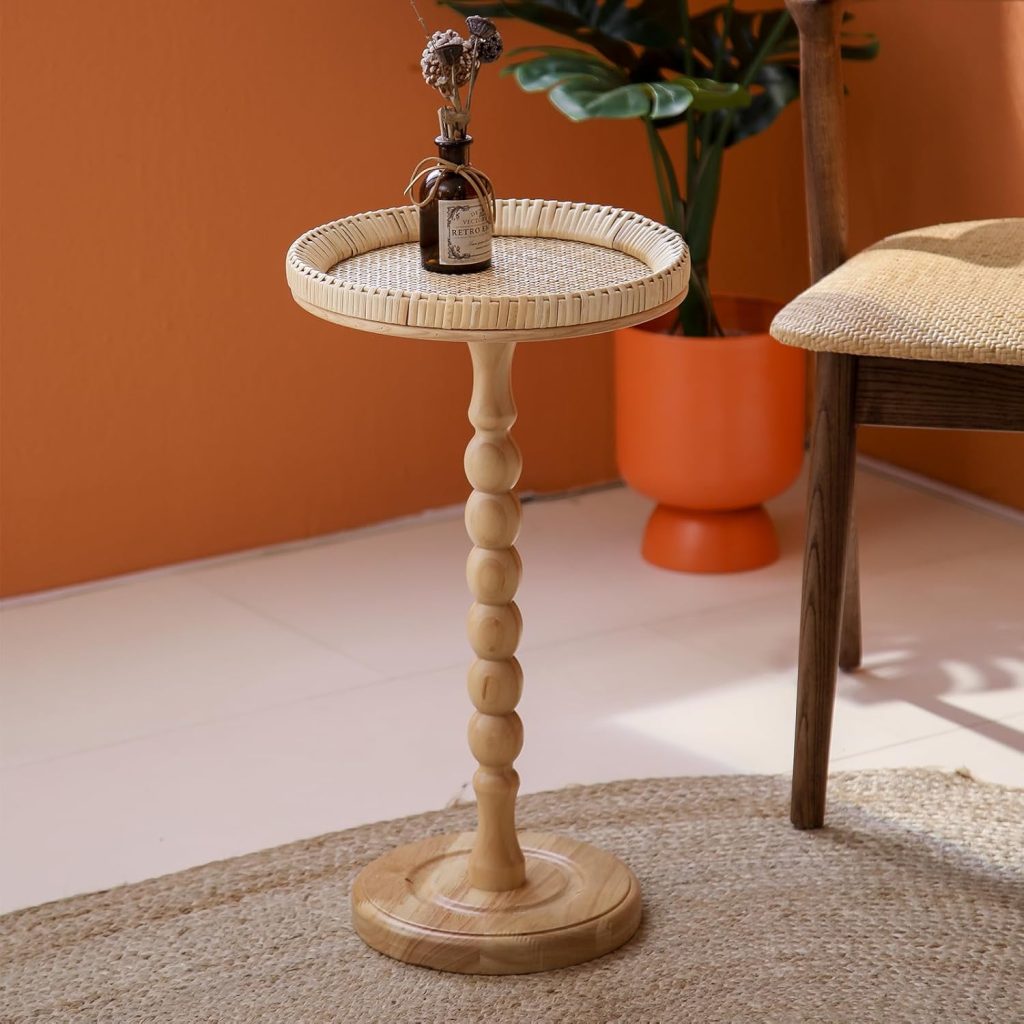 A small round wooden side table with a beaded pedestal base holds a small brown vase with dried flowers. Nearby, a woven chair and a potted plant complement the warm, organic look—perfect for pairing with rattan furniture to create an inviting natural space.