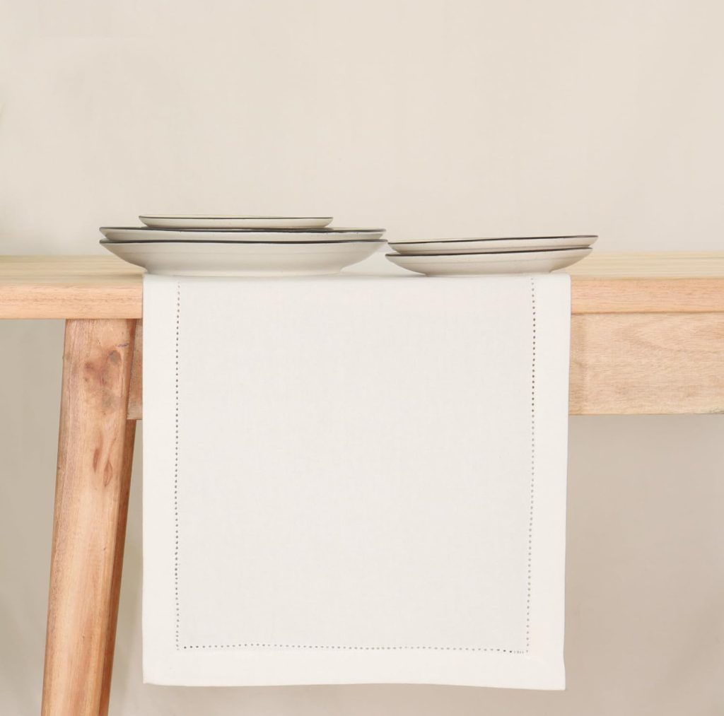 Four stacked white plates sit on a wooden table with a white table runner hanging down the side.