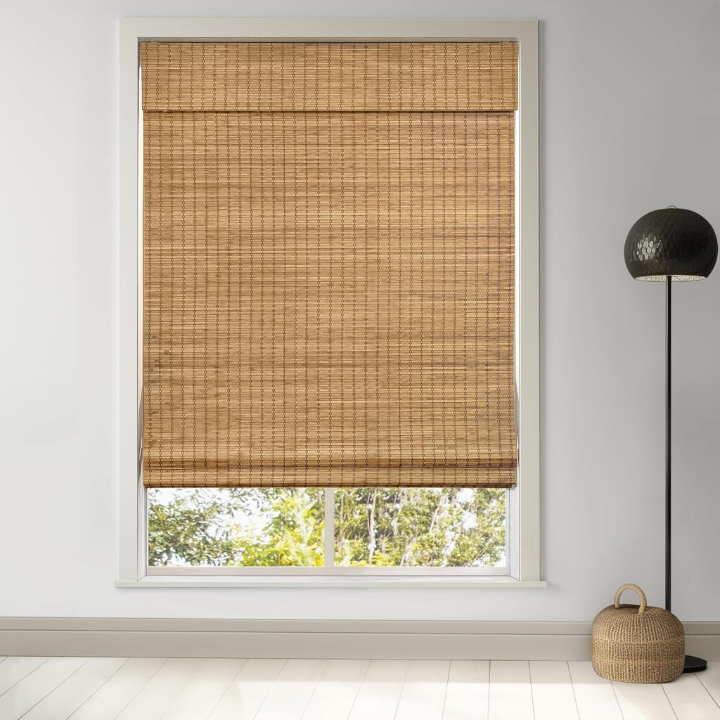 A window with a rolled-down bamboo shade, a black floor lamp, and a round wicker basket on a light wood floor create an inviting scene. The touch of greenery visible through the lower part of the window complements the natural textures and hints at the presence of rattan furniture nearby, enhancing the warm and organic atmosphere.