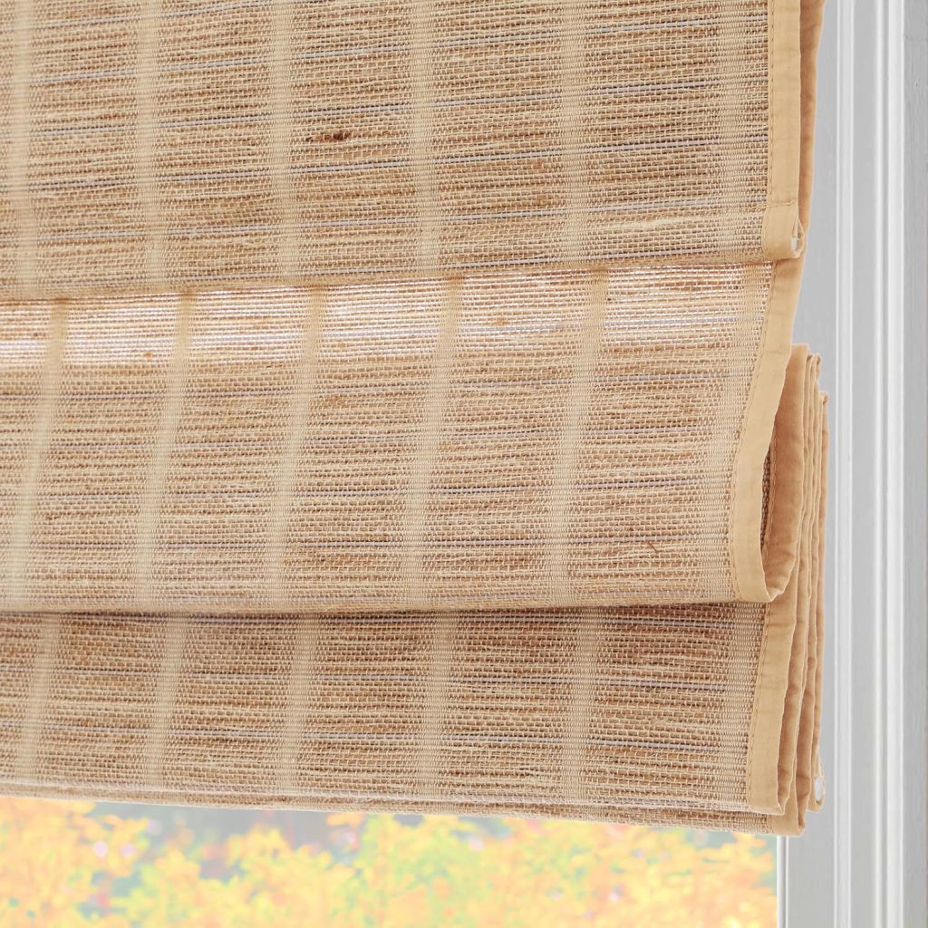 Close-up of a beige woven Roman shade, partially raised, hanging in front of a window with blurred autumn foliage visible outside—an inviting scene that pairs beautifully with rattan furniture for a warm, natural aesthetic.