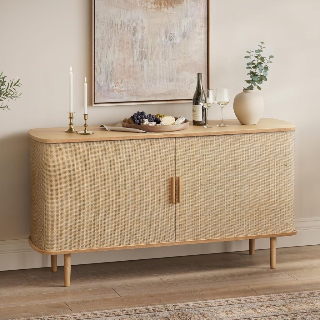 A light wood sideboard with rounded edges, styled to complement rattan furniture, is topped with a tray of fruit and cheese, two wine glasses, a bottle, candlesticks, and a vase with greenery. A painting hangs above.