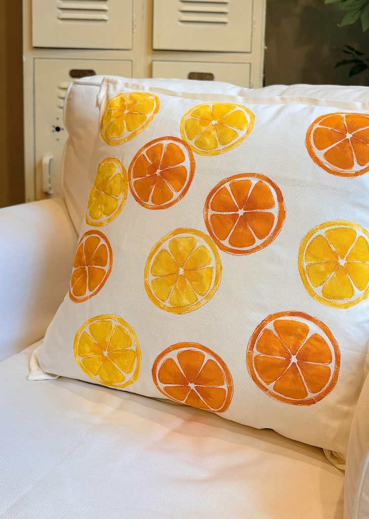 White throw pillow with orange and yellow citrus slice designs resting on a white couch, with lockers and a plant blurred in the background.