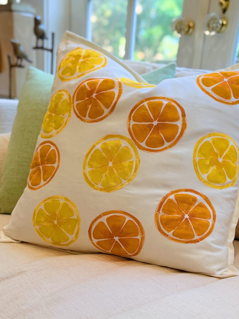White pillow with orange and yellow citrus slice designs sits on a light-colored couch with a green pillow and window in the background.