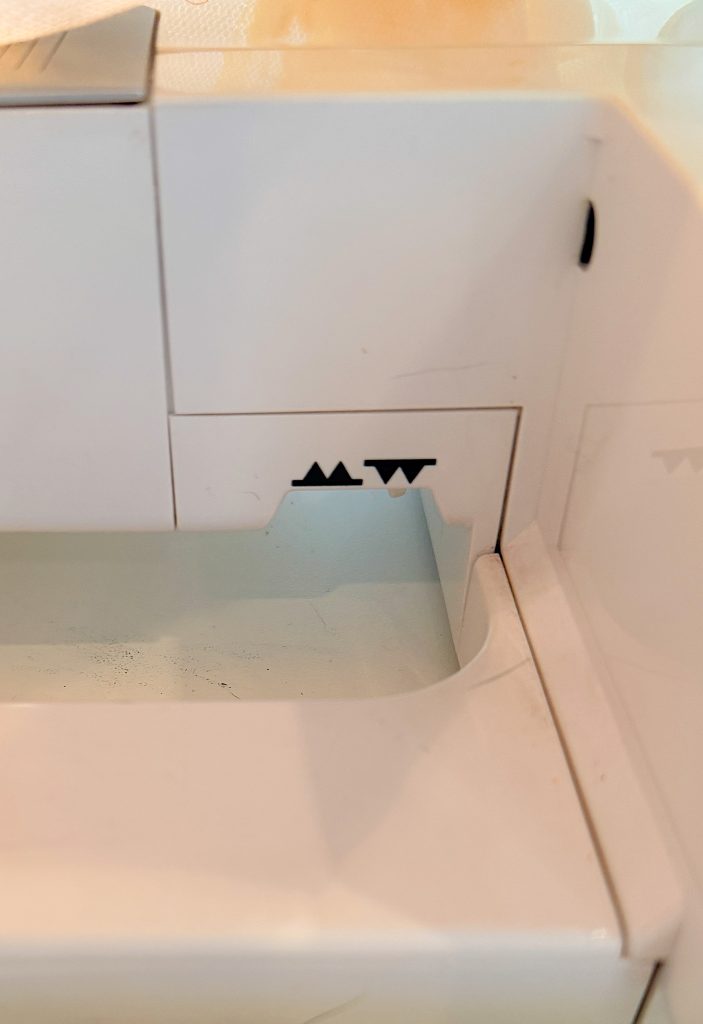 Close-up of a white appliance with black zigzag and straight line symbols printed above a small compartment.