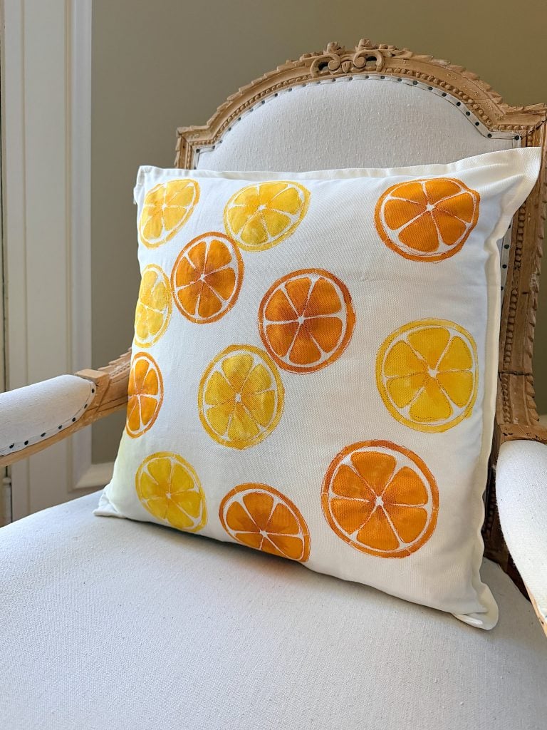 A white throw pillow with orange and yellow citrus slice prints rests on a light-colored upholstered wooden chair.