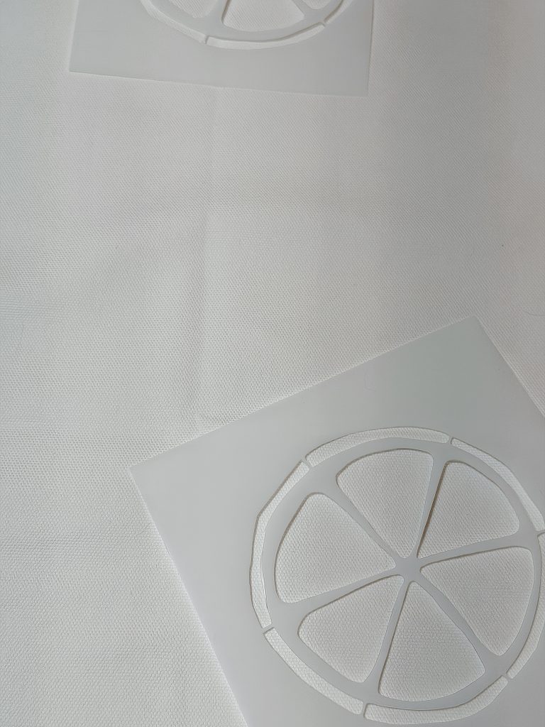 Two white sheets with citrus fruit slice cutout designs are placed on a textured white surface, with one in the foreground and one partially visible in the background.