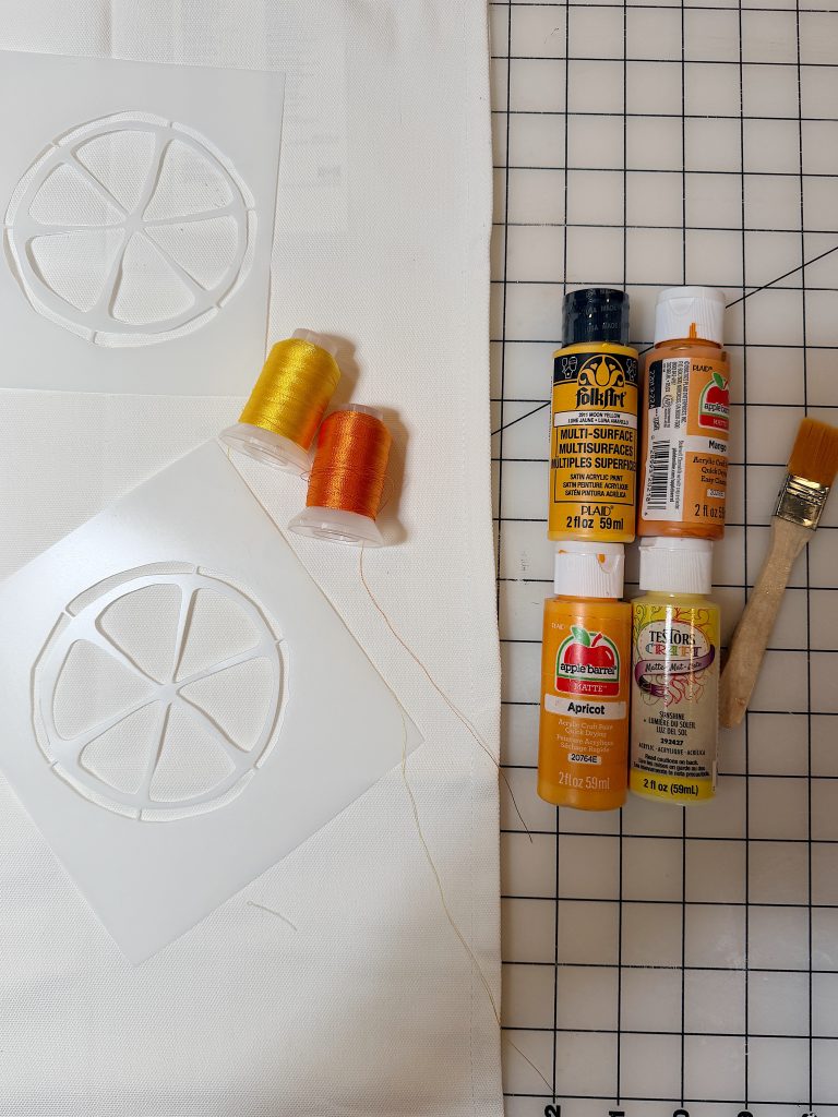 Two citrus slice stencils, yellow and orange thread spools, four bottles of orange-yellow acrylic paint, and a paintbrush on white fabric over a grid cutting mat.