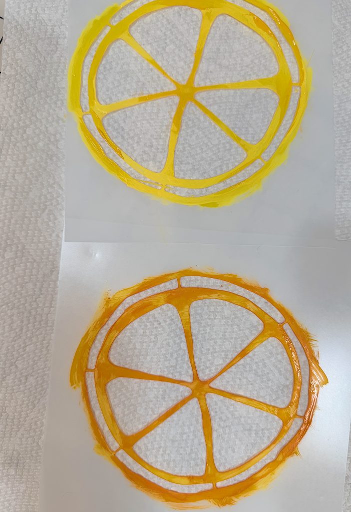 Two circular shapes resembling citrus fruit slices, one yellow and one orange, are painted on sheets of white paper towels.
