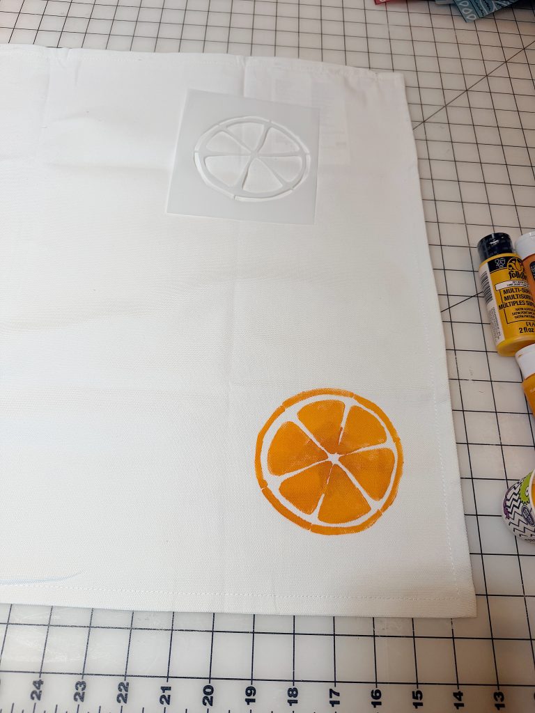 A white cloth with a painted orange slice and a matching stencil nearby, placed on a grid cutting mat. Yellow and orange paint bottles are to the right.