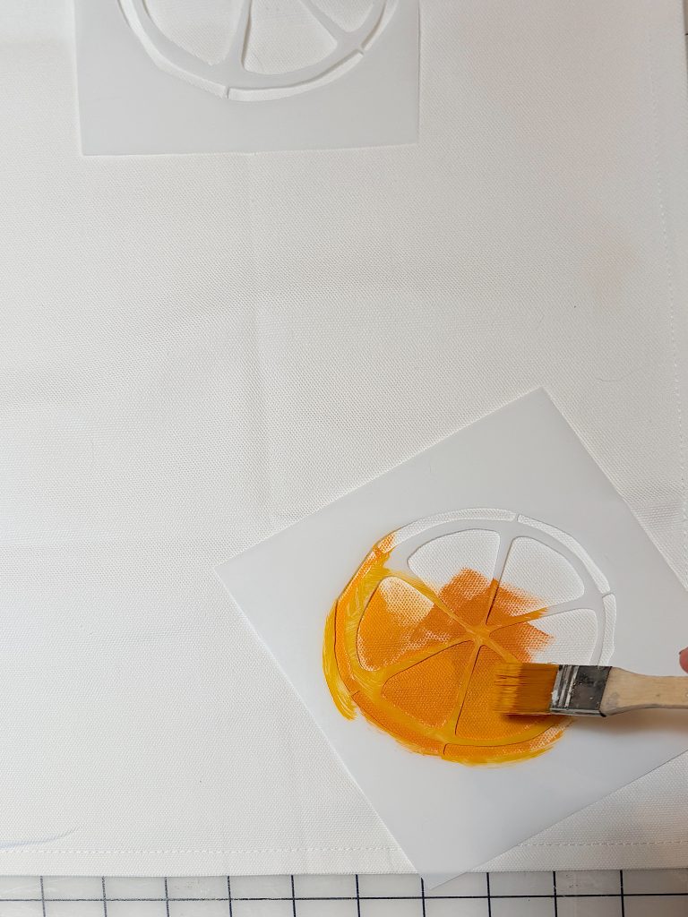 A hand uses a paintbrush to apply orange paint to a stencil of a citrus fruit slice on white fabric.