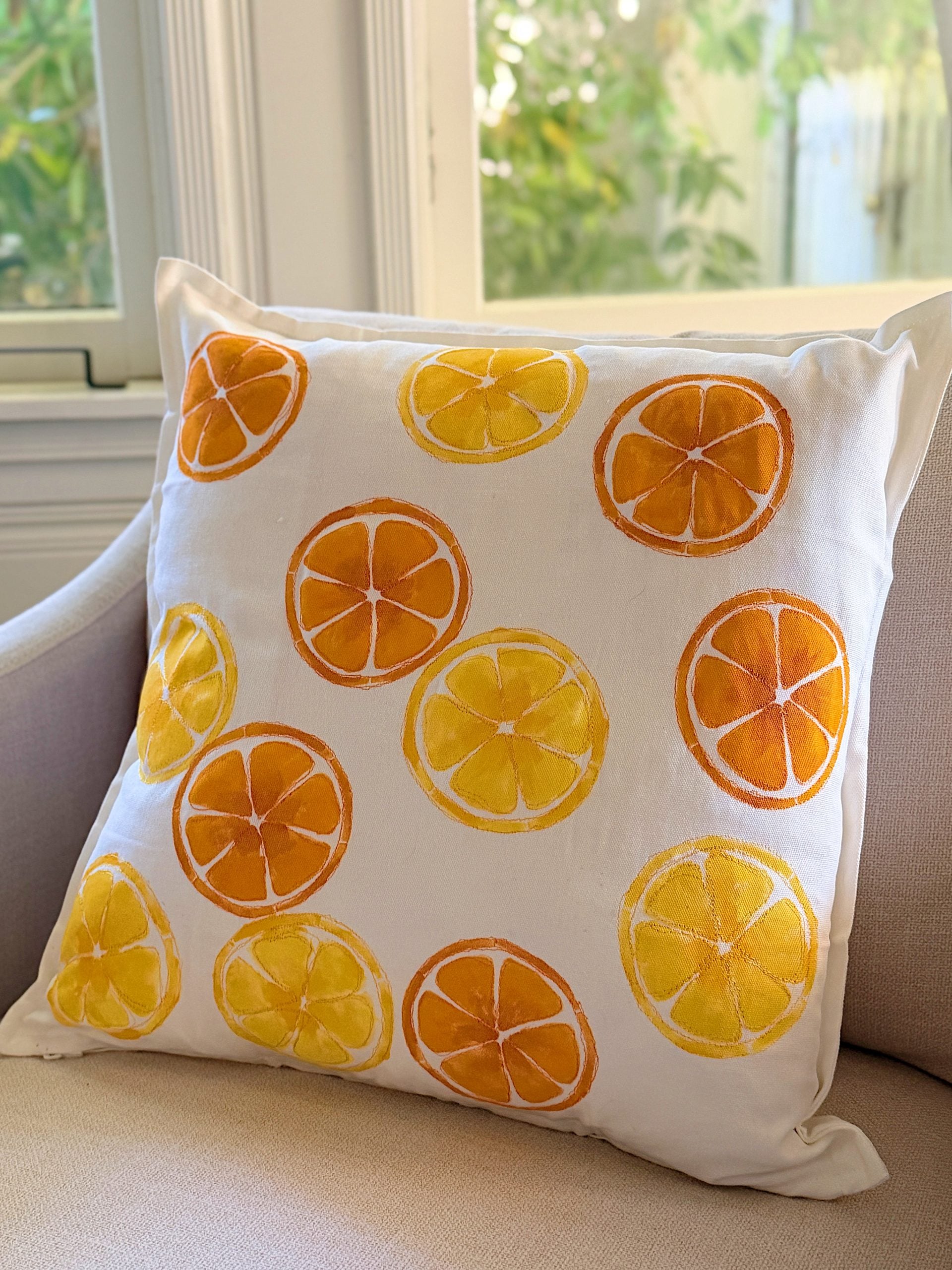 A white throw pillow with orange and yellow citrus fruit slice designs sits on a beige armchair near a window.