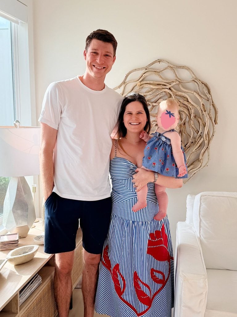 A man and woman stand indoors by a white couch; the woman holds a baby wearing a blue dress. The group smiles at the camera amid bright, natural light and neutral decor.
