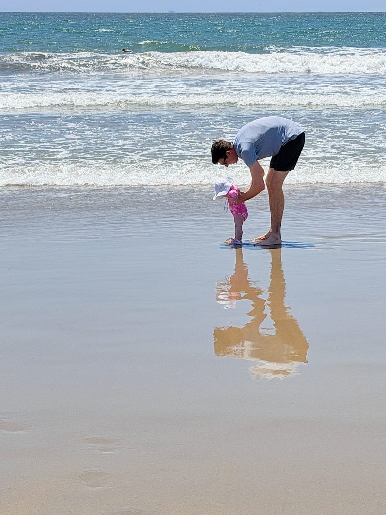 An adult bends down to help a small child standing at the edge of the ocean on a sandy beach, their reflections visible in the wet sand.