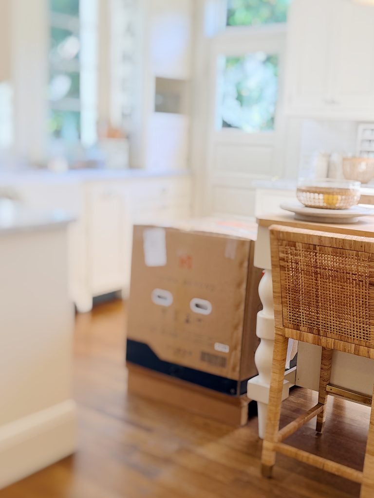A cardboard box is placed on the floor next to a kitchen island with a woven chair in a bright, modern kitchen.