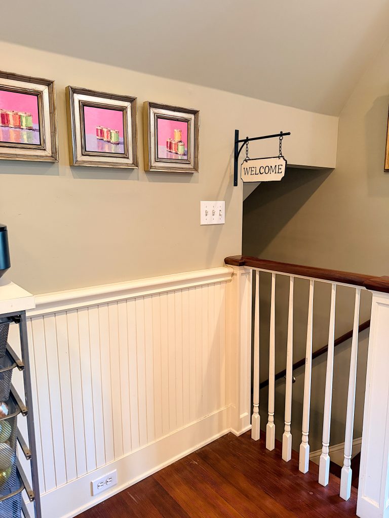 A staircase with a wooden banister, cream wainscoting, three framed pictures, and a hanging "Welcome" sign on the wall.