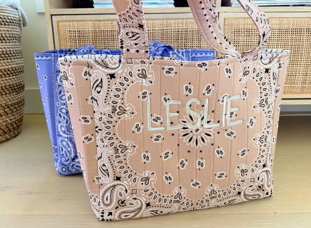 A pink tote bag with a paisley bandana pattern and the name "LESLIE" stitched on the front sits in front of a blue bag with a similar pattern.