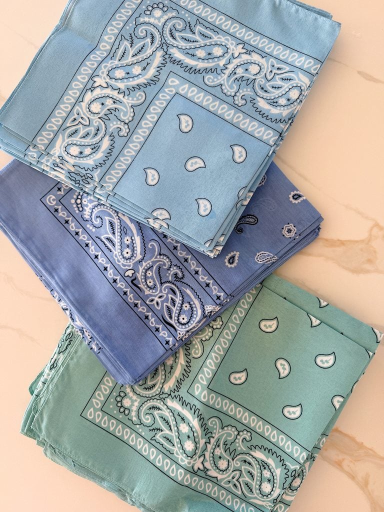 Three folded bandanas in light blue, blue, and green with paisley patterns are stacked on a marble surface.