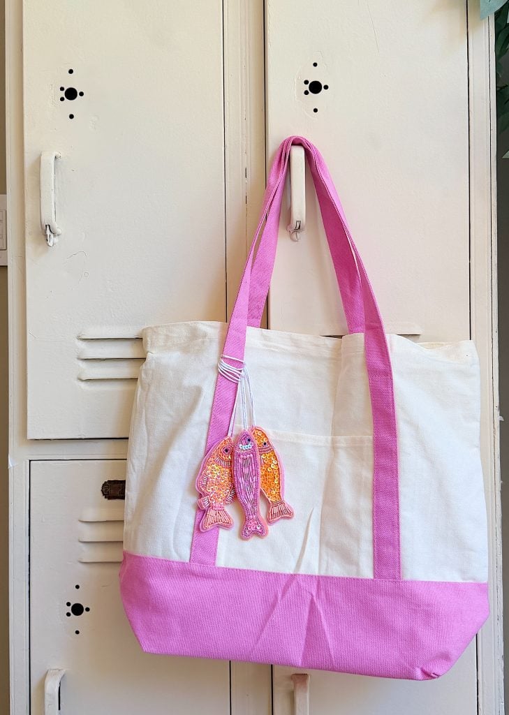 A white and pink tote bag with pink handles hangs on a locker. A sequined fish keychain is attached to one handle.