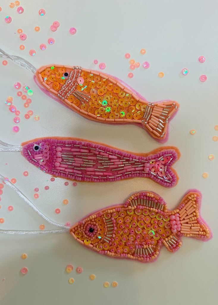 Three beaded fish ornaments in shades of orange and pink lie on a white surface, surrounded by scattered small round sequins.