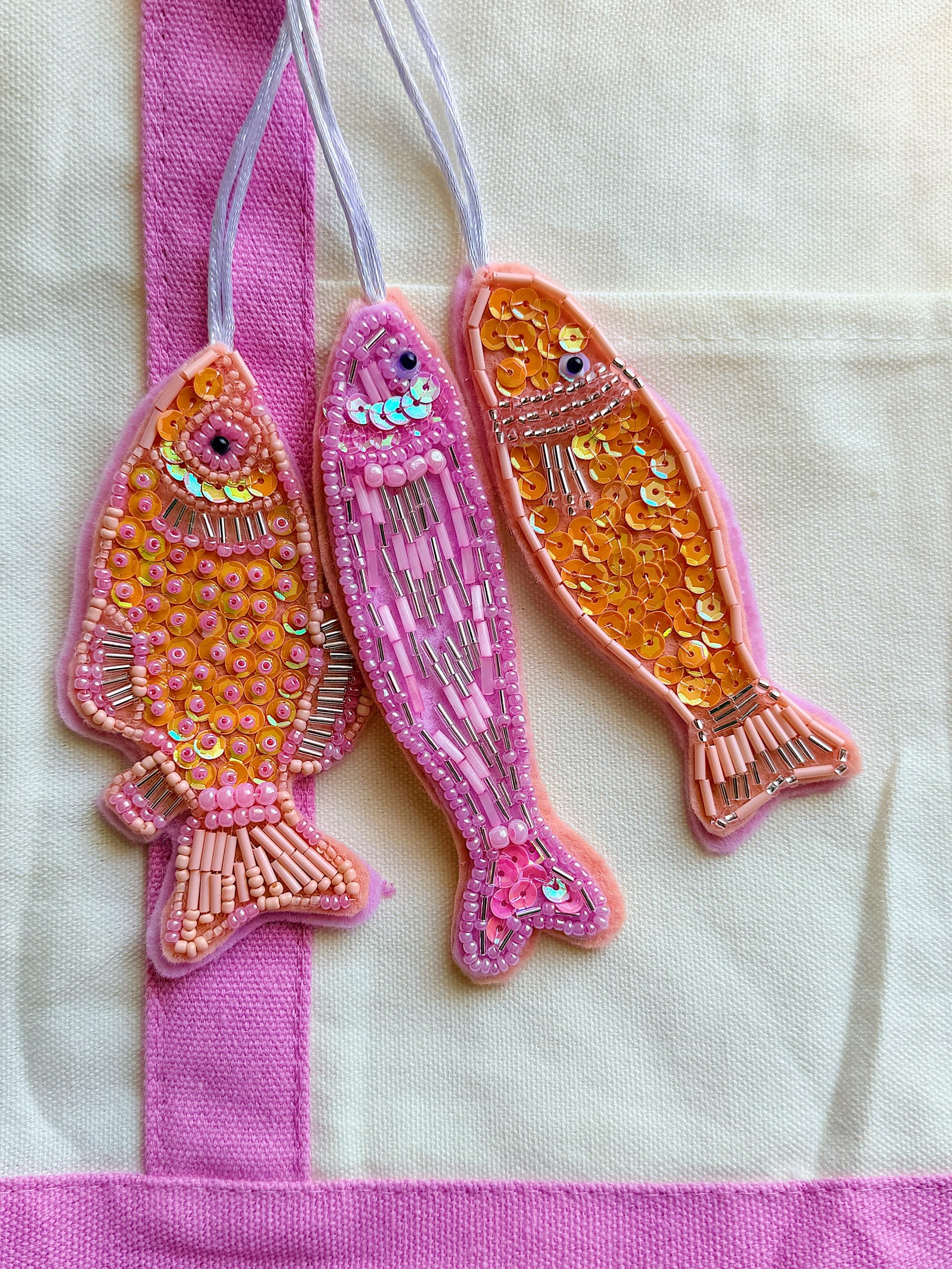 Three pink and orange fish-shaped ornaments decorated with sequins and beads are displayed on a cream and pink fabric background.