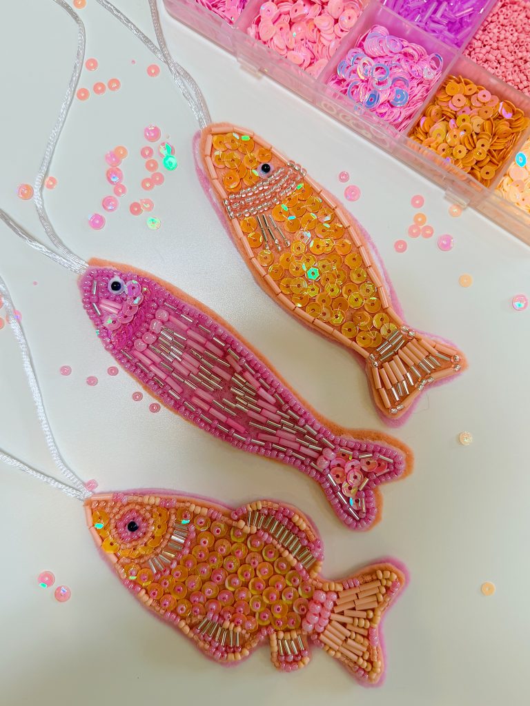 Three beaded fish-shaped ornaments in pink and orange tones are displayed on a white surface, with a box of matching sequins and beads in the background.