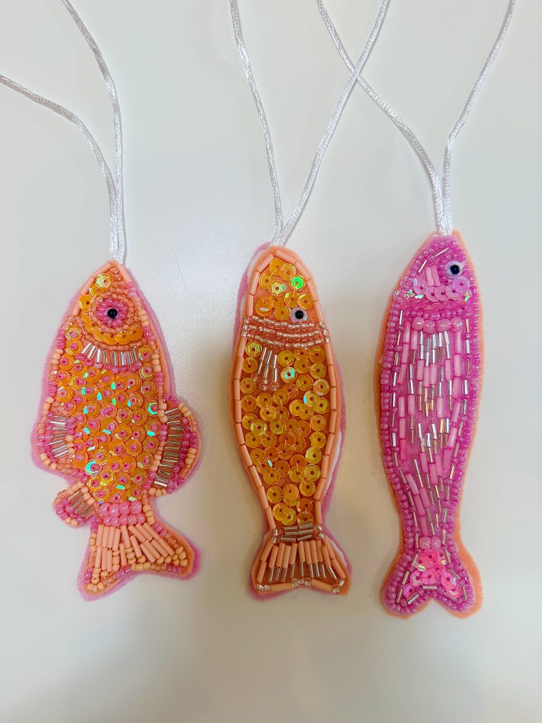 Three pink and orange fish-shaped ornaments decorated with sequins and beads hang by silver threads against a white background.