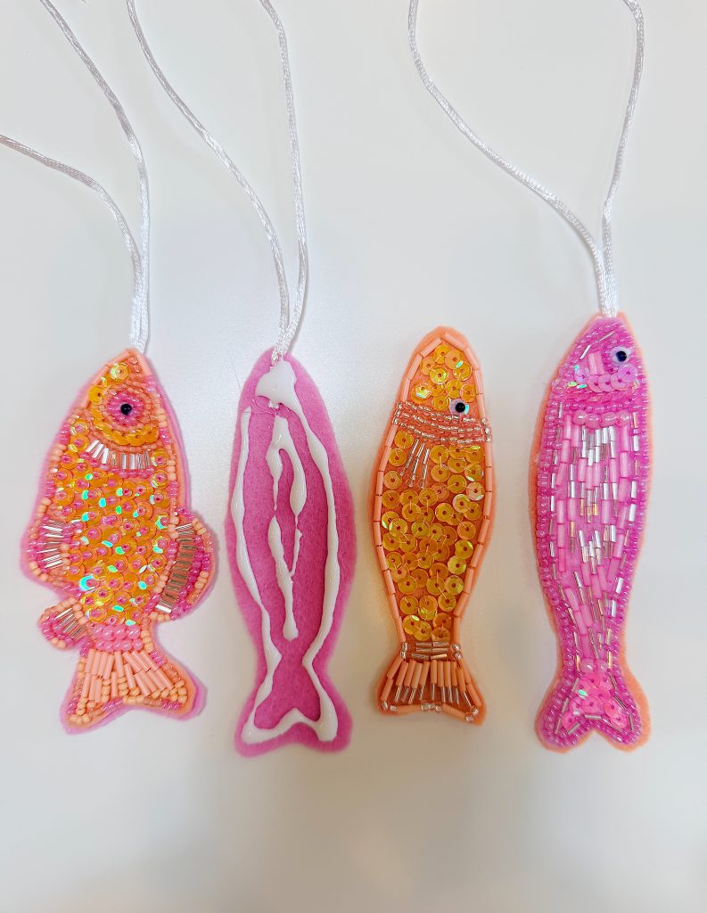 Four fish-shaped ornaments hang by white cords; three are decorated with beads and sequins, and one features pink and white felt. All are arranged vertically on a white background.