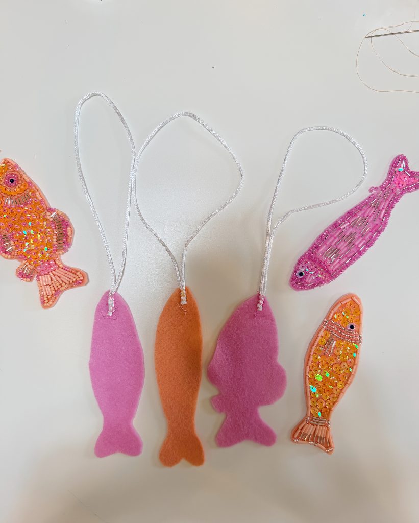 Five fish-shaped ornaments on a white surface; three are plain felt with strings, two are decorated with sequins and embroidered designs.