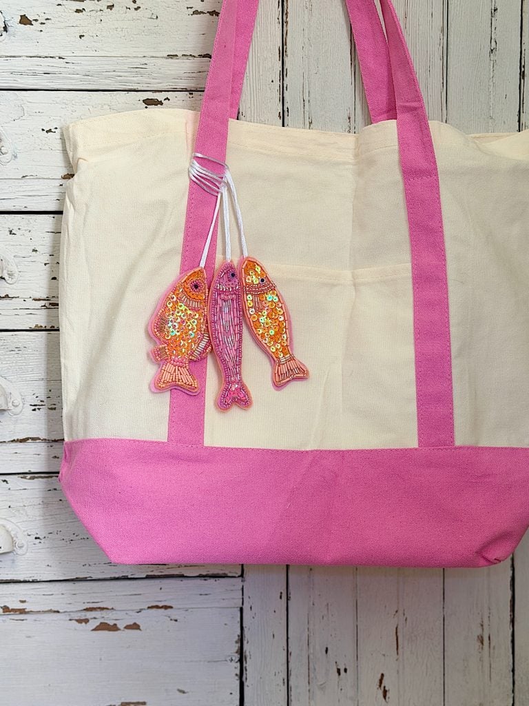 A beige and pink tote bag hangs on a white wooden wall, decorated with three colorful fish-shaped keychains attached to one handle.