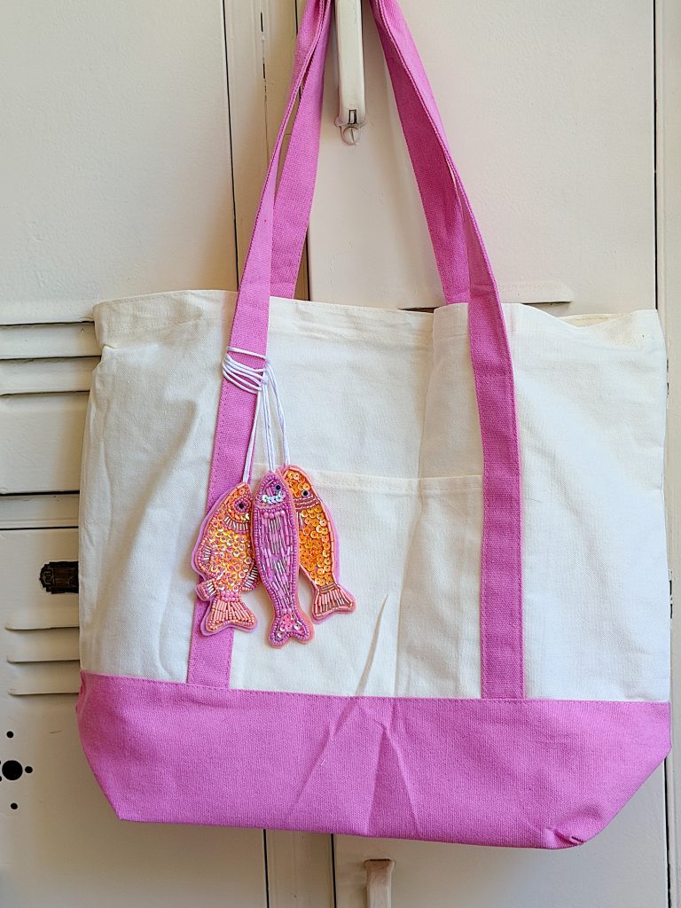 A white and pink tote bag hangs on a hook, decorated with a keychain featuring three sequined fish.