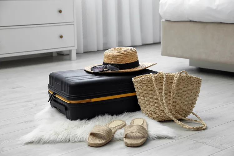 A closed black suitcase on a white rug with a straw hat, sunglasses, woven bag, and straw sandals next to it in a bedroom with light flooring.