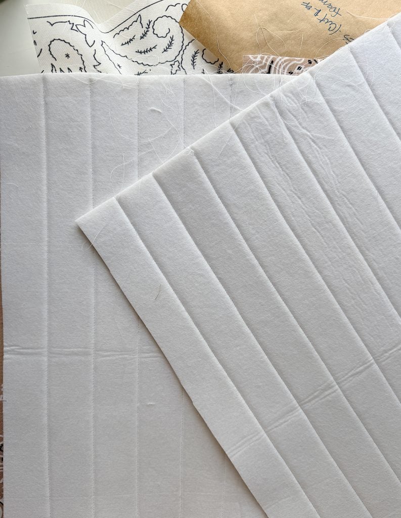 Two pieces of white quilt batting are laid out, with a brown paper sewing pattern and black-and-white printed fabric partially visible underneath.