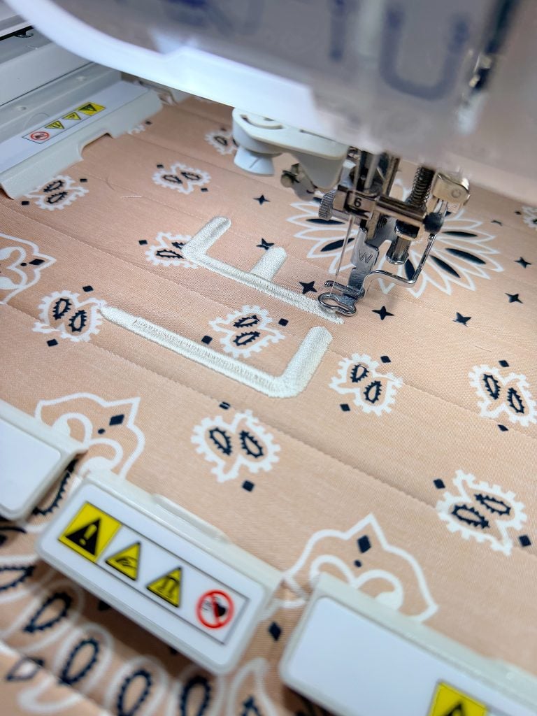 Close-up of an embroidery machine stitching a white letter "L" onto patterned beige fabric with floral and geometric designs.