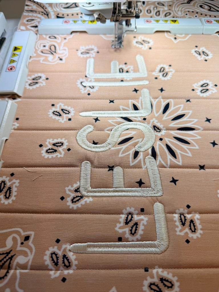 A sewing machine embroiders the word "lush" in white thread on a peach fabric with a floral and paisley pattern.