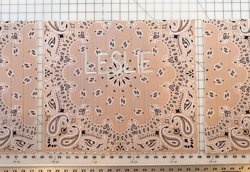A light brown bandana with a white paisley pattern has the name "LESLIE" embroidered in white letters in the center, placed on a grid cutting mat with rulers.