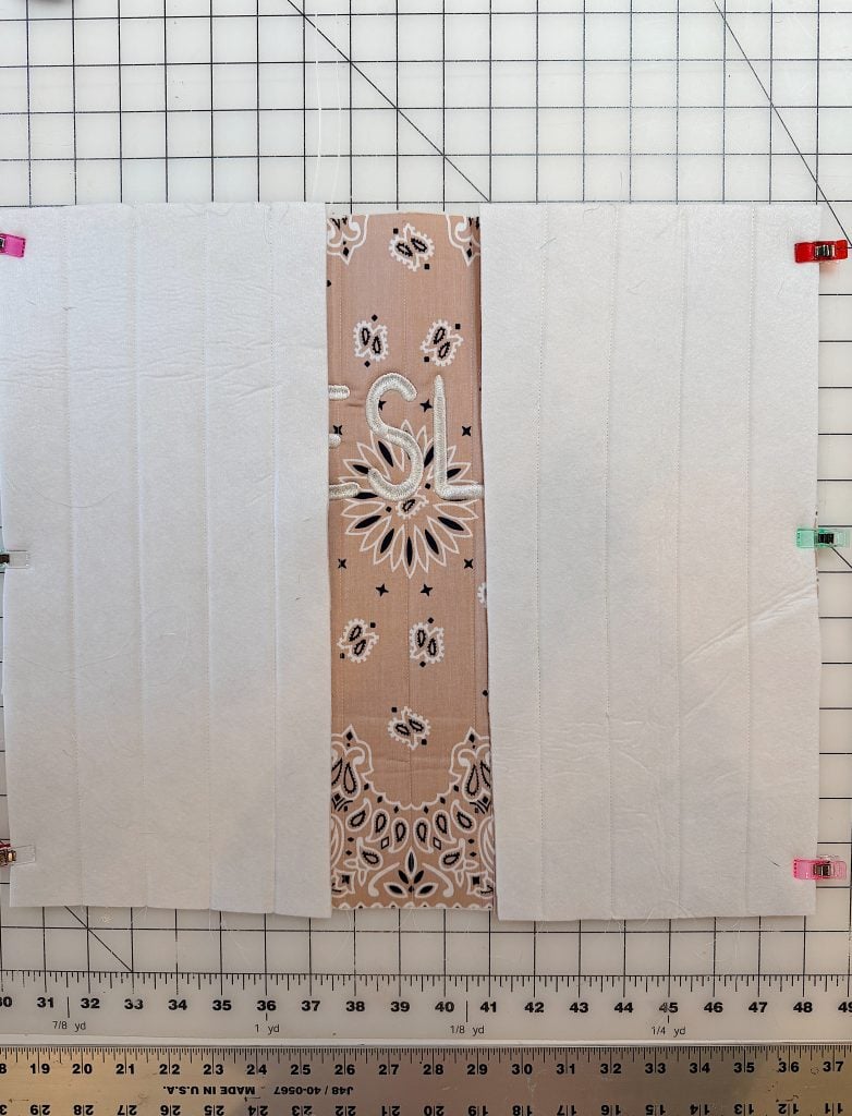 A piece of fabric with a patterned center strip and solid white sides is laid flat on a gridded cutting mat, secured with colorful clips along the edges.