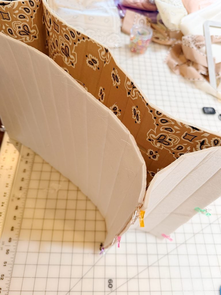 Partially constructed garment piece with beige fabric on the outside and patterned fabric lining, held together by clips, on a grid cutting mat.