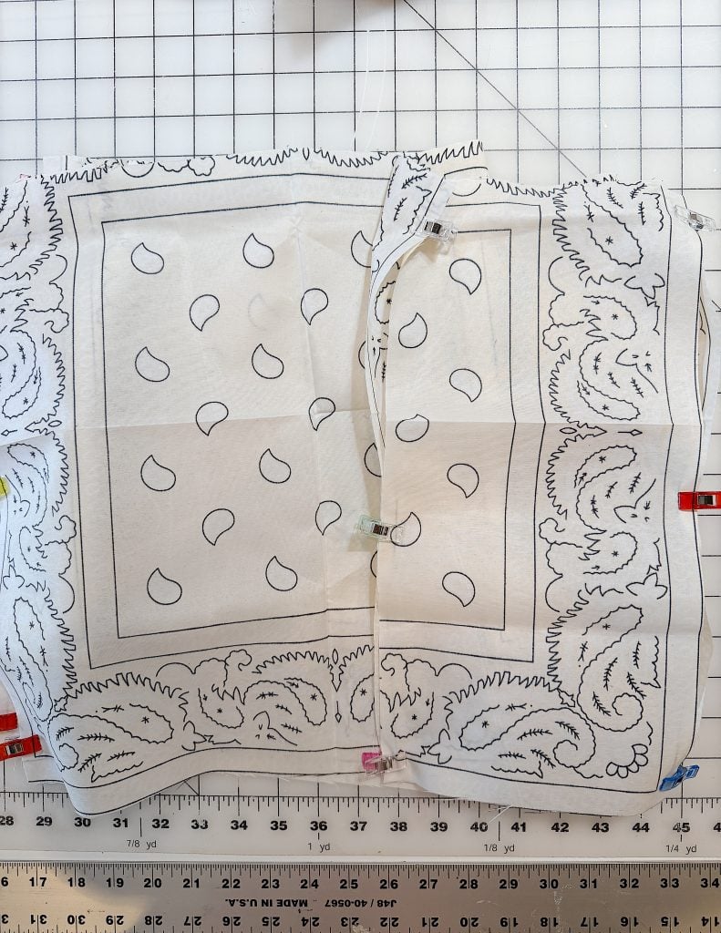 A patterned bandana fabric is laid out on a gridded cutting mat with measuring marks, held in place by sewing clips along the edges.