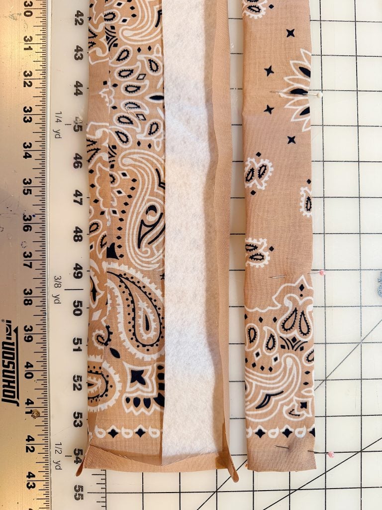 Two strips of paisley-patterned fabric, one partially folded with interfacing, are placed on a cutting mat next to a ruler marked in inches.