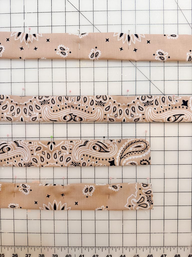 Four strips of tan bandana fabric with black and white patterns are laid out on a gridded cutting mat with pins along the edges. A ruler is visible at the bottom.
