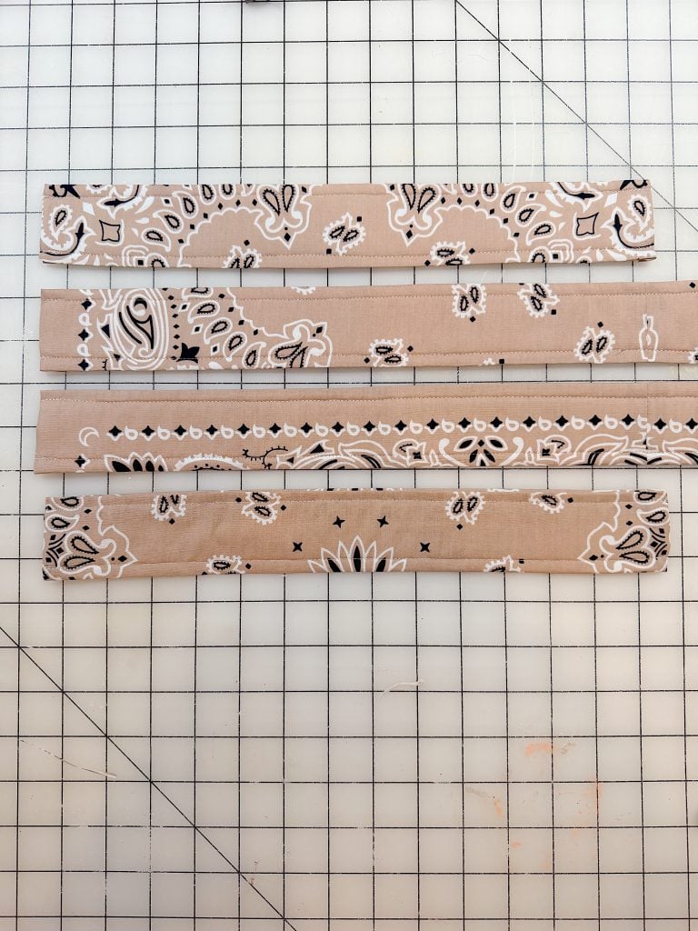 Four rectangular strips of cardboard with a tan and white paisley bandana pattern, laid out horizontally on a gridded cutting mat.