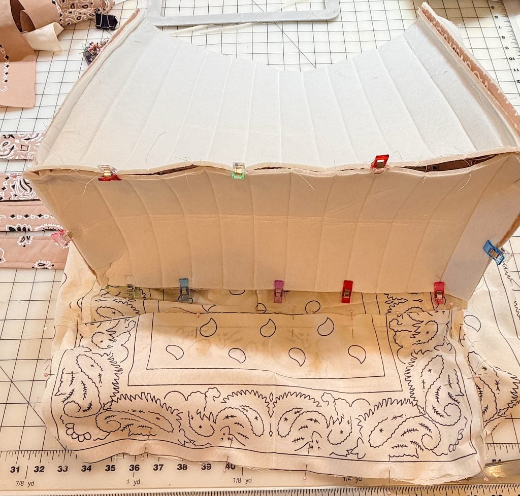 A partially assembled corset with fabric clips attaches cream-colored panels to a patterned cloth, laid out on a gridded cutting mat with sewing supplies nearby.