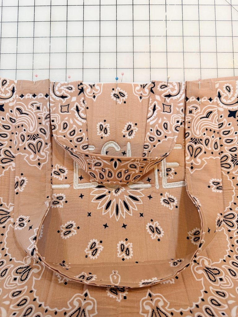 A peach-colored bandana fabric tote bag with a front pocket is displayed on a grid-patterned cutting mat.