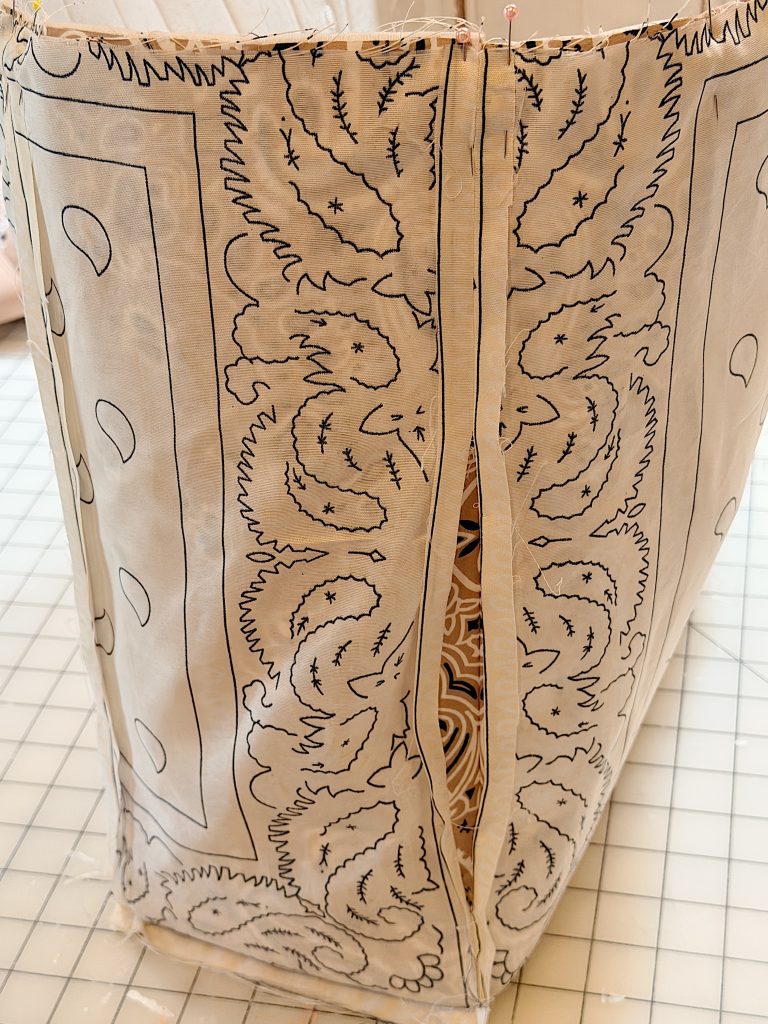 A piece of fabric with a black paisley design is sewn into a rectangular shape, with an open seam held by pins on a gridded cutting mat.