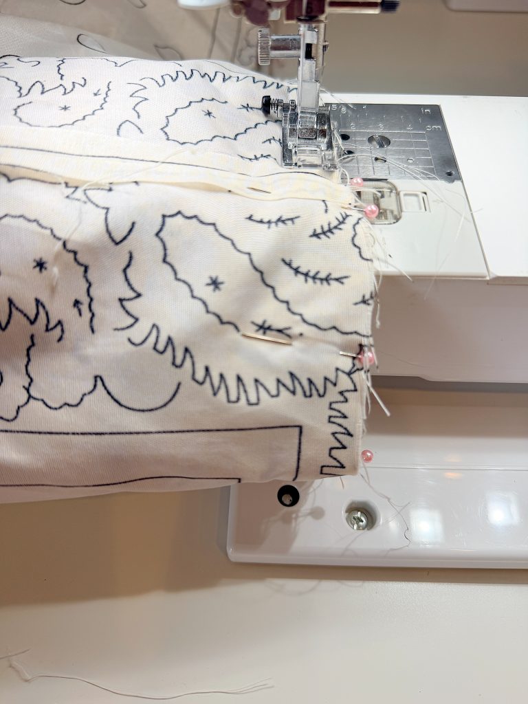 Close-up of fabric with a black line pattern being sewn together on a sewing machine, with pins holding the fabric in place.